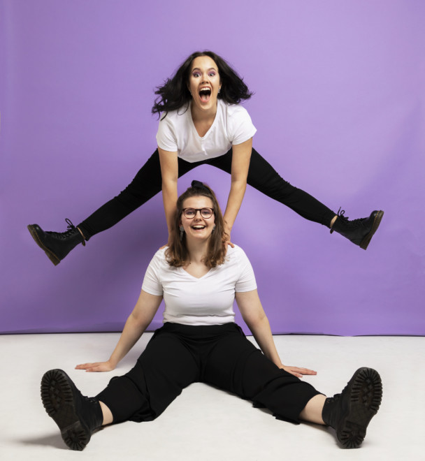 Kaksi nuorta naista, joista toinen istuu lattialla ja toinen on takana ja hyppää ilmaan jalat auki. Violetti tausta ja valkoinen lattia.