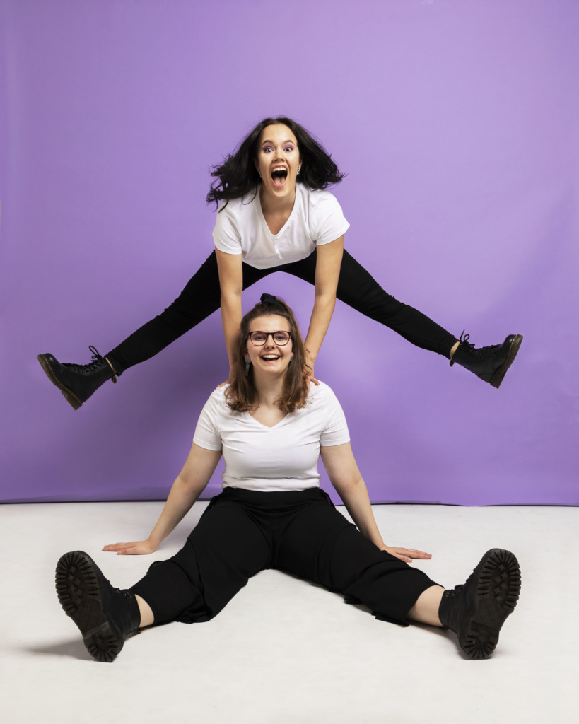 One girl sitting on the floor and other one behind her jumping with legs spread . Violet background.