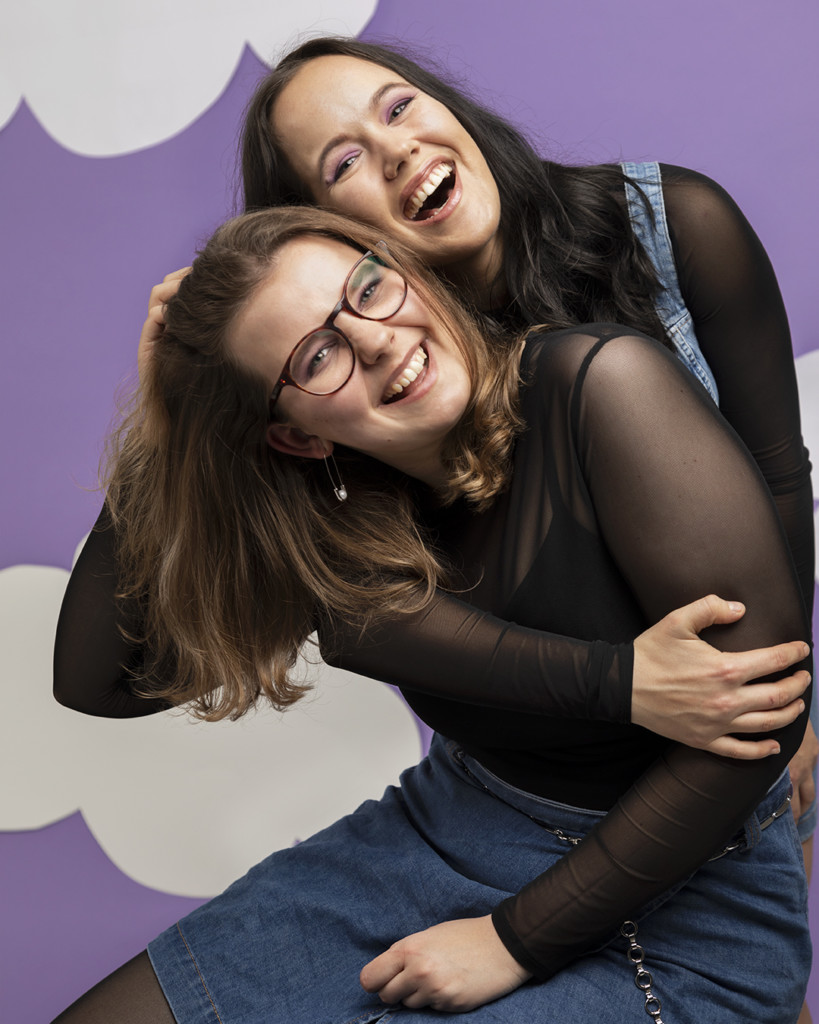 Two girls sitting on the floor and hugging. Violet background with white clouds. 