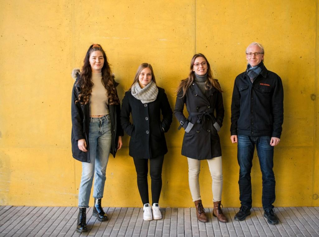 Murrost team of four standing outside, in front of a yellow wall