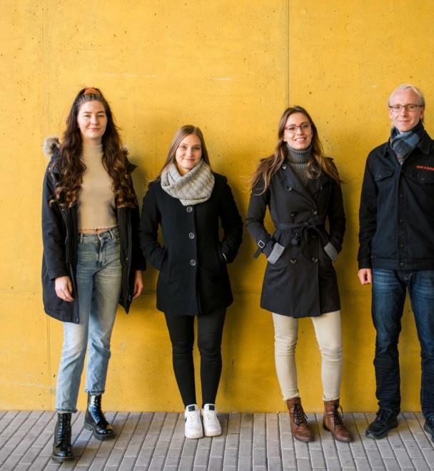 Murrost team of four standing outside, in front of a yellow wall