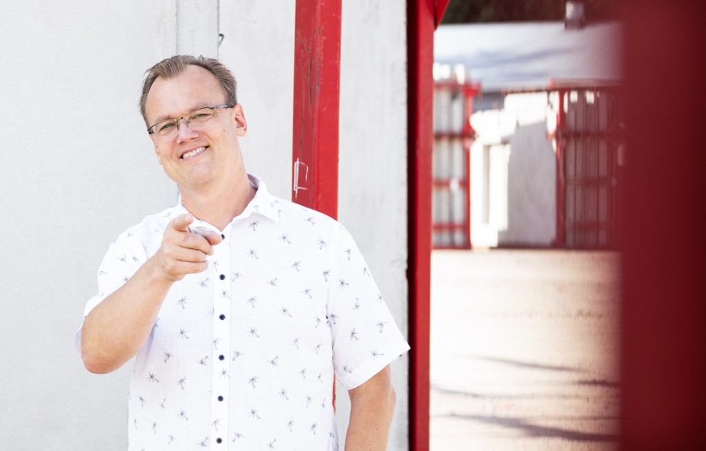 Antti Leijala standing in front of a white wall, pointing to the camera and smiling. Red details in the background.