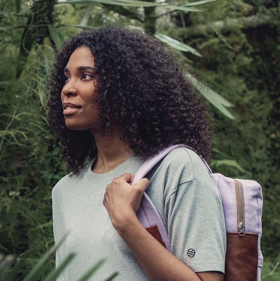 A woman in grey t-shirt carrying a back bag on her left shoulder. Green plants on the background.