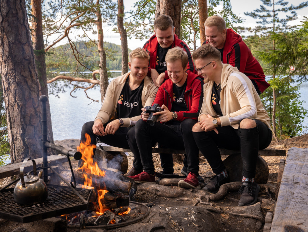 Three men sitting by a camp fire, the middle one holding a camera on his hands. Two more men standing behind the three men. All looking on to the camera screen.