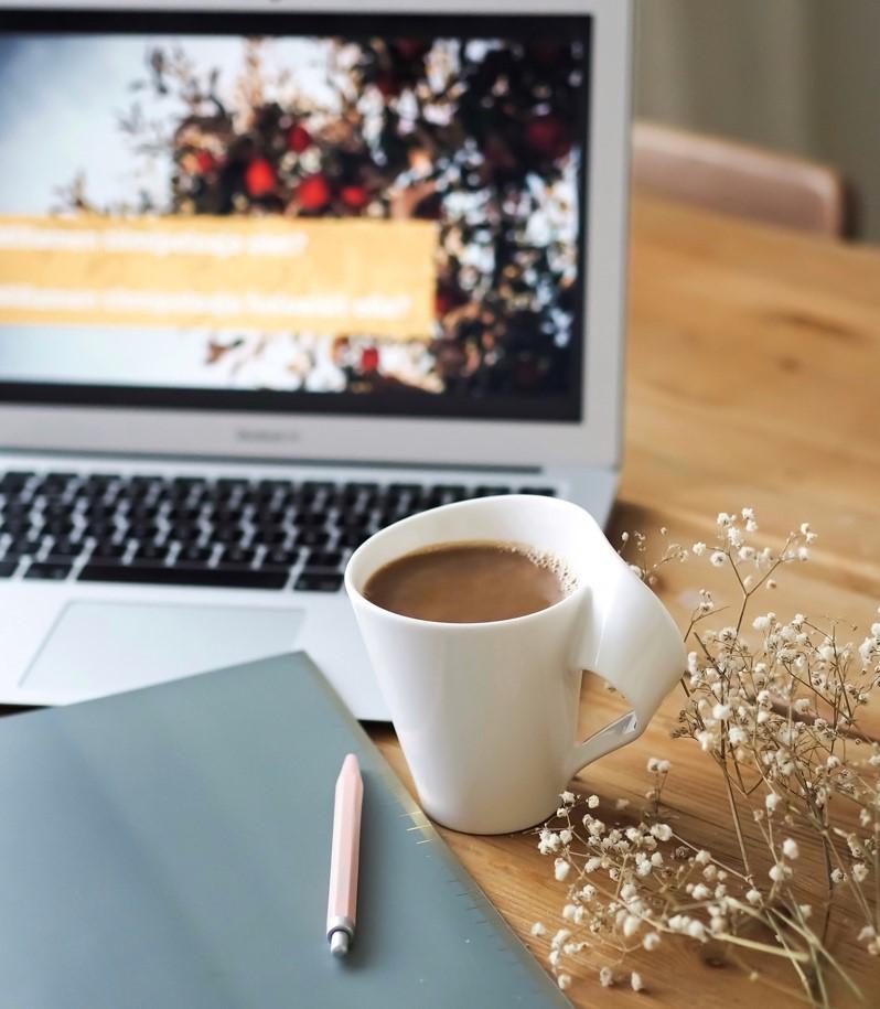 A notebook and a pen, a cup of coffee in a white mug, open laptop on the background.