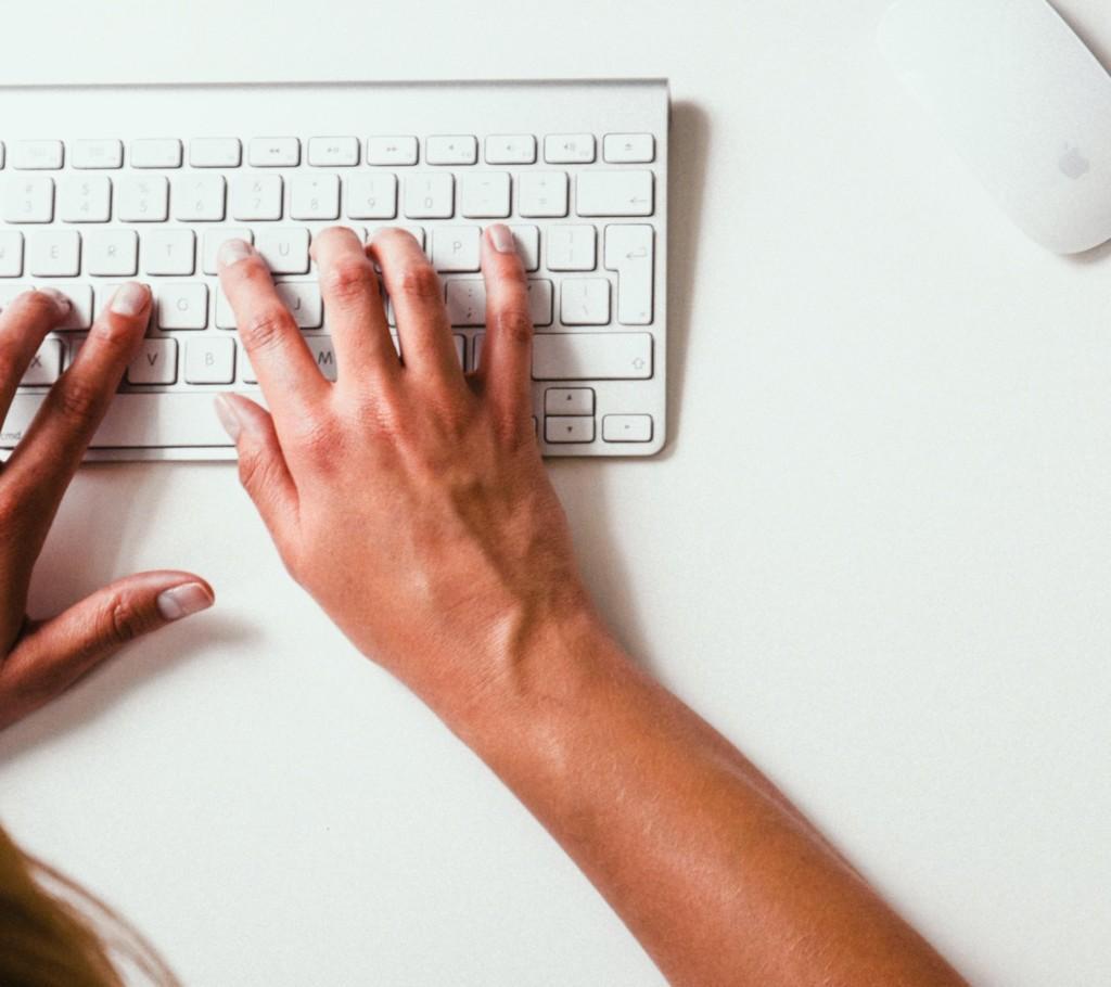 Photo taken from above; hands on white keyboard. A yellow watch on the right side of the table.