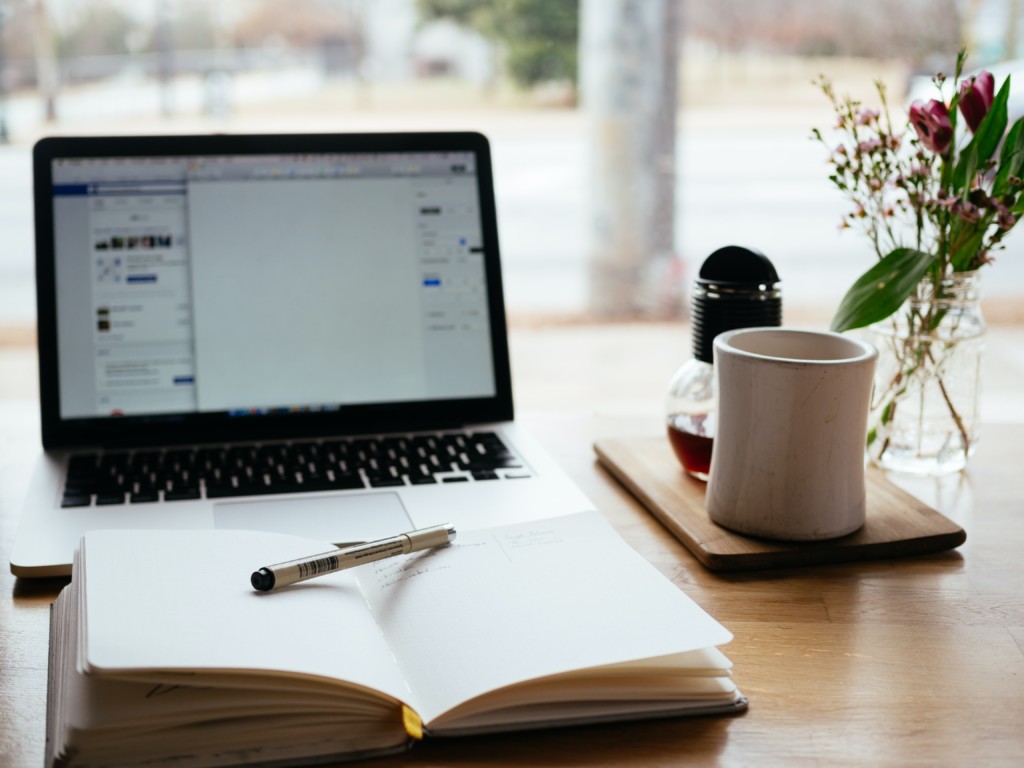 A desk in front of a window, an open laptop,  a coffee mug and an open notebook with a pencil on it.