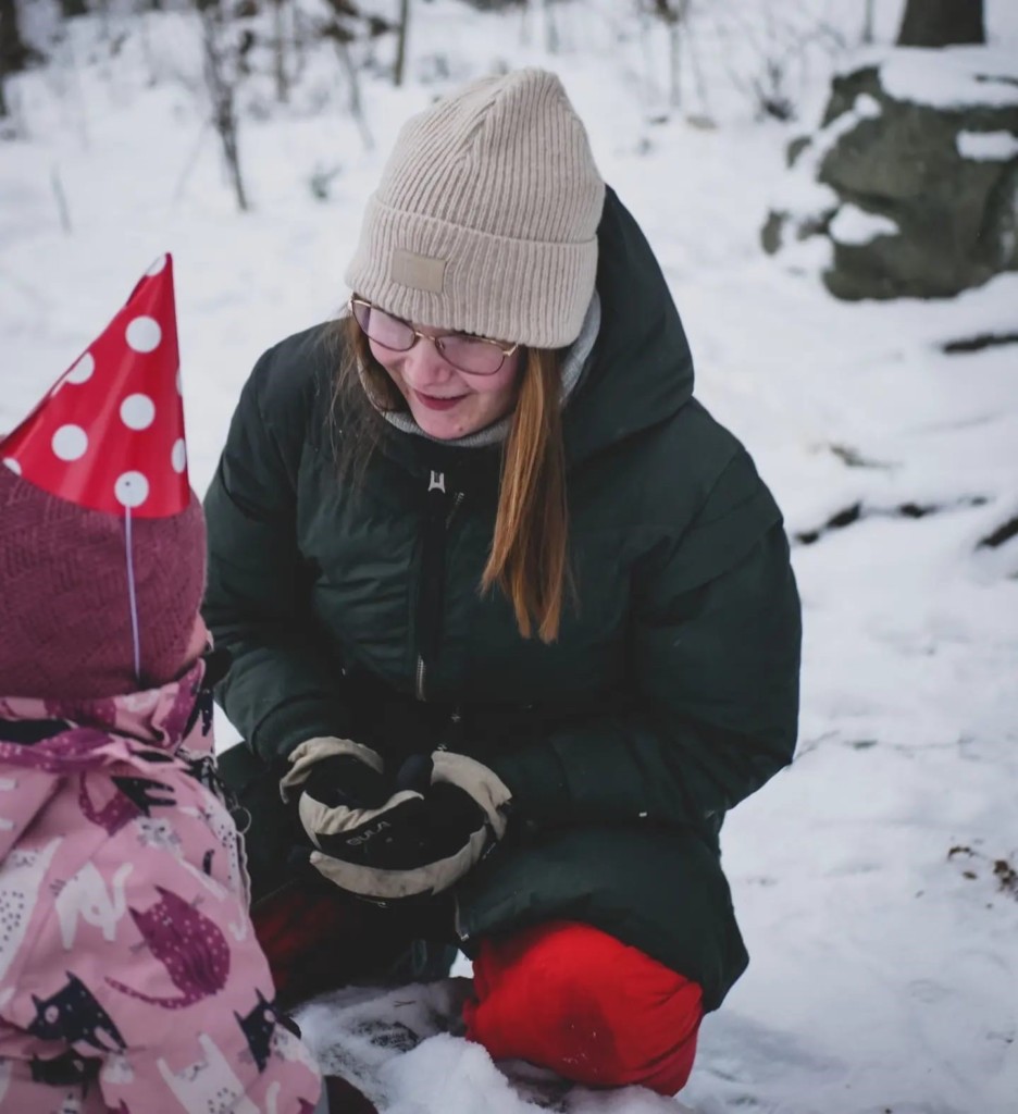 A woman on her knees in the snow, talking to a toddler with a red triangle hat on. 