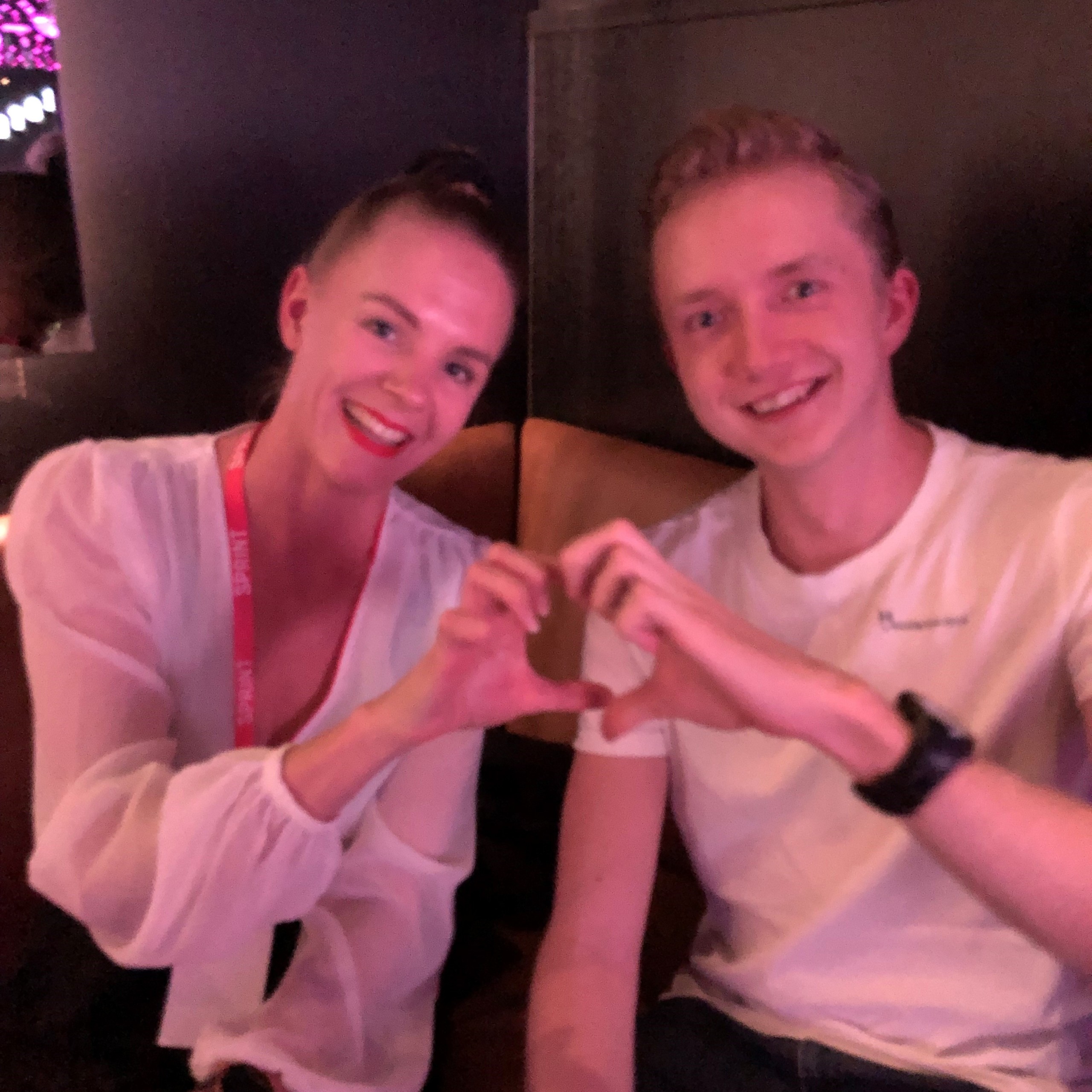 Two people sitting next to each other forming a heart with their fingers.