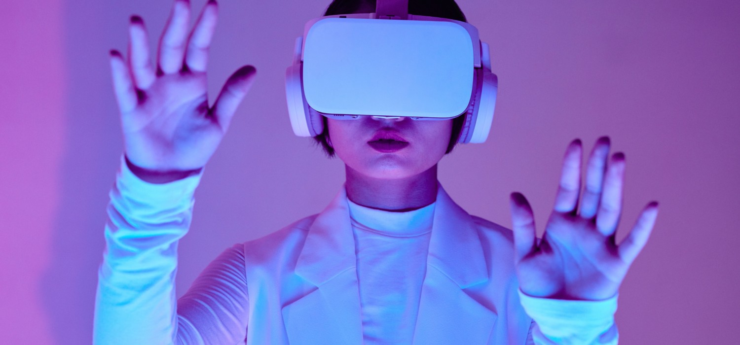 Valkoisiin pukeutunut henkilö, jolla on virtuaalilasit päässään, koskettaa ilmaa edessään kämmenet eteenpäin. Kuvan värimaailma on pastellissävyillä valaistu.