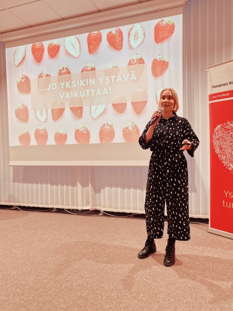 Saara giving a presentation at a Red Cross event. She is wearing a white-spotted pantsuit.