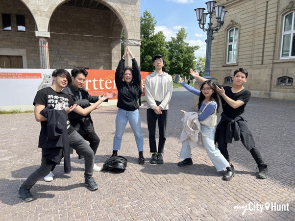 Six people standing in a half circle. Old buildings in the back ground.