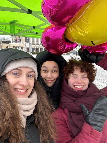 Three people smiling and standing outside in a tent, one person holding a colourful flower shaped balloon.